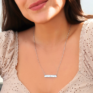 A close-up of a woman wearing a delicate silver bar necklace with a small engraved design, paired with a beige eyelet top.