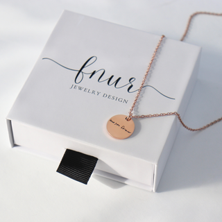 A rose gold necklace with a round engraved pendant reading 'may you Dream' draped over a white jewelry box branded 'fnur JEWELRY DESIGN'.