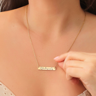 A close-up of a woman wearing a delicate gold necklace with a horizontal bar engraved with a blue heartbeat line and small symbols. A hand is gently touching the pendant.