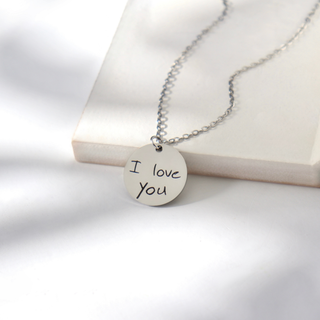 A silver round pendant necklace with 'I love you' hand-stamped on the front, resting on a white display block against a white background.