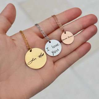 Three round engraved pendant necklaces in gold, silver, and rose gold tones are displayed on a person's hand. The gold pendant reads 'Jane, Mom', the silver pendant reads 'I love you', and the rose gold pendant reads 'love you forever'.