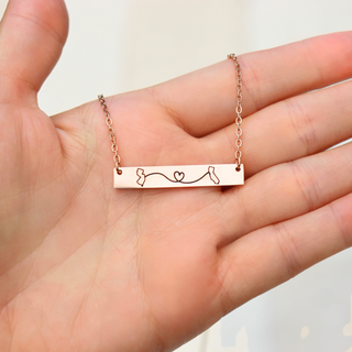 A rose gold bar necklace with a delicate chain and engraved baby bootie design, held in the center of an open hand.