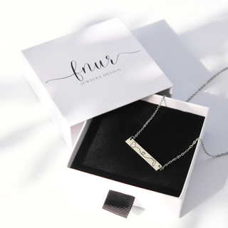 A silver bar necklace with engraved cursive script is displayed in an open white jewelry box with a black velvet insert. The brand name 'fnur JEWELRY DESIGN' is printed on the lid.