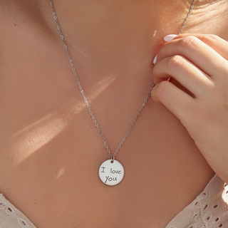 A close-up lifestyle shot of a silver chain necklace with a round silver pendant engraved with 'I love You' in black script, worn by a model.