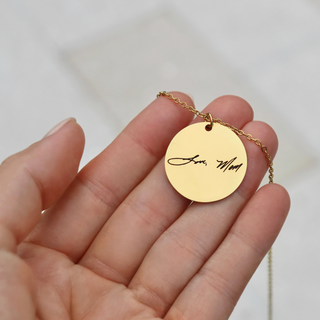 A gold round pendant necklace engraved with 'Love, Mom' in black script, held in a person's hand against a soft white background.