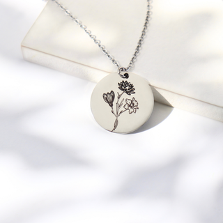 A silver chain necklace with a round white pendant engraved with black floral illustrations, displayed on a white textured surface with soft leaf shadows.