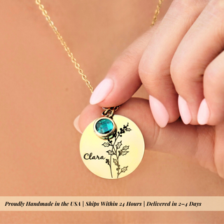 A close-up of a hand with pale pink manicured nails holding a gold pendant necklace. The pendant is round and engraved with the name 'Clara' and a delicate floral design, topped with a small round turquoise-blue gemstone.