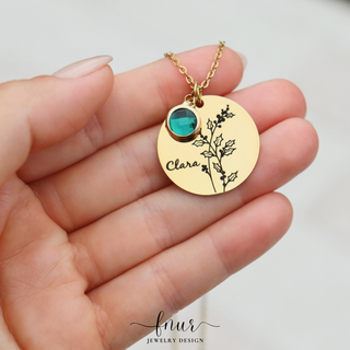 A gold round pendant necklace with a black botanical engraving and the name 'Clara', held in a hand. The pendant features a small round turquoise-blue stone.